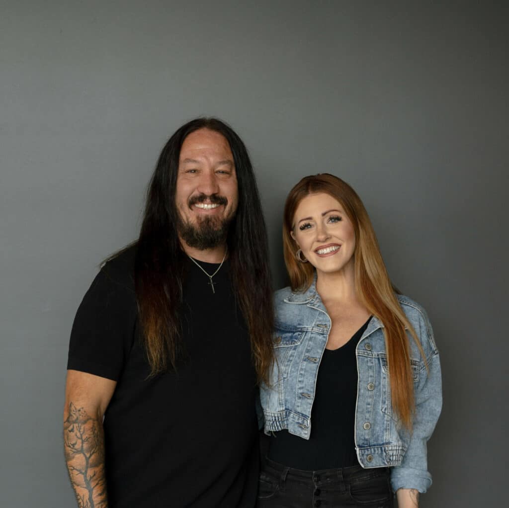 A man with long dark hair and a black t-shirt stands next to a woman with long light brown hair, wearing a black top and a denim jacket. They are posed together against a gray background.
