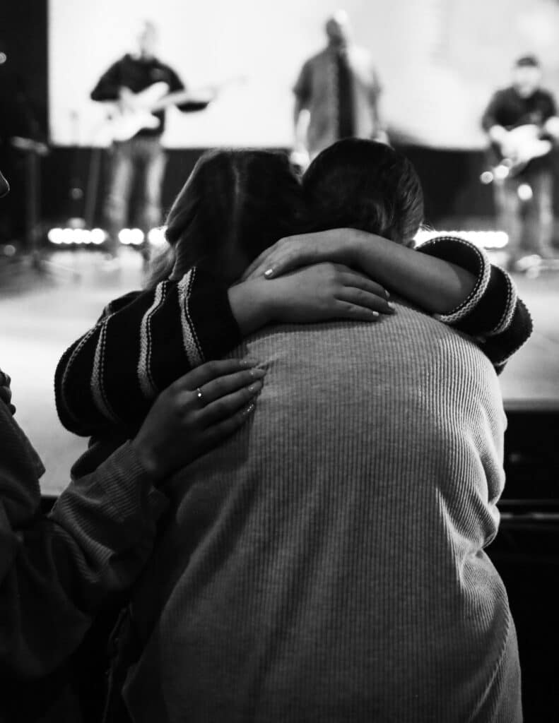 Two individuals embrace each other while watching a band perform on stage. The image is in black and white, with soft lighting creating a warm atmosphere, and band members playing instruments in the background.