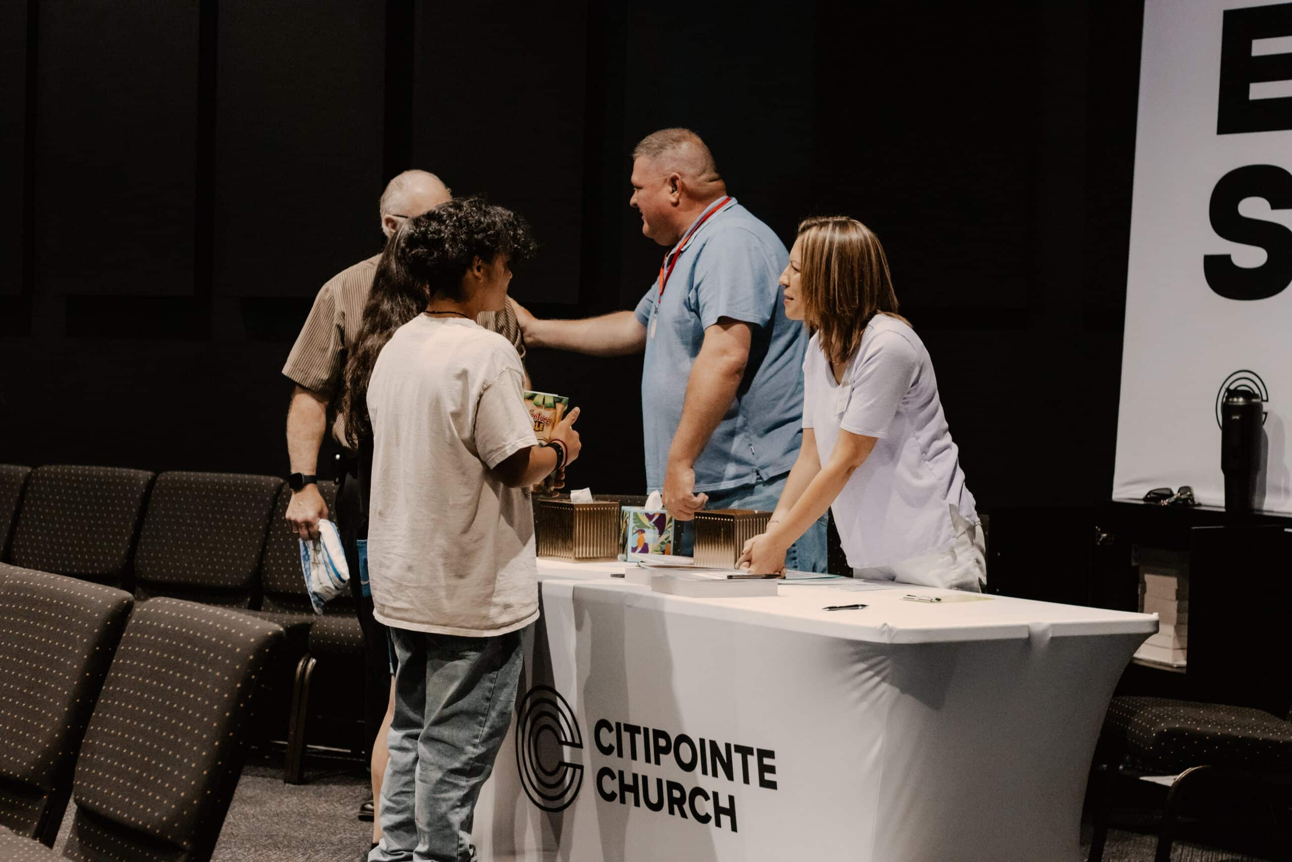 A group of people interacting at a table in a church setting. Two individuals are standing behind the table, while two others are engaged in conversation, with seating visible in the background. The table has a Citipointe Church logo.