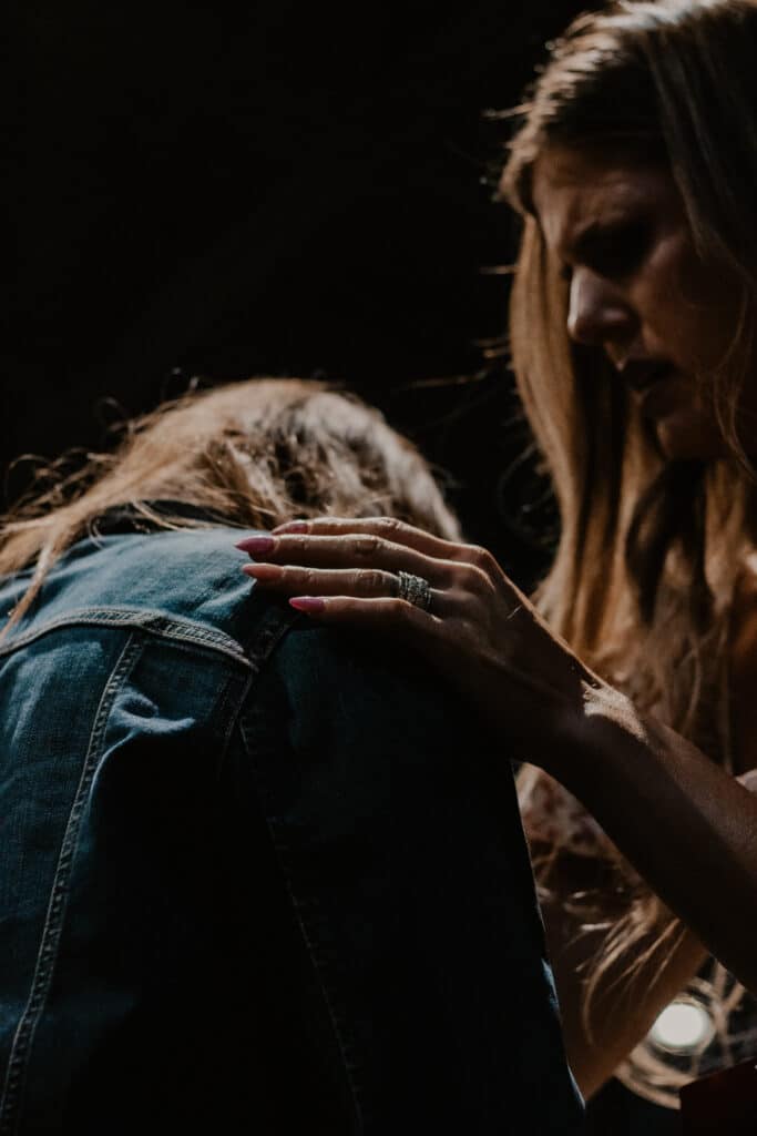A woman with long hair reaches out to touch the shoulder of another person who is facing down, partially obscured by long hair and wearing a denim jacket. The setting is dimly lit with a focus on their hands.