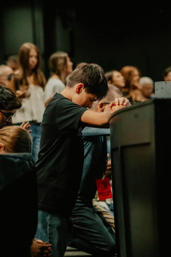 A young boy with dark hair wearing a black shirt kneels beside a seated person, resting his arms on a dark surface in front of him. In the background, several people are standing, and the setting appears to be a gathering or service.