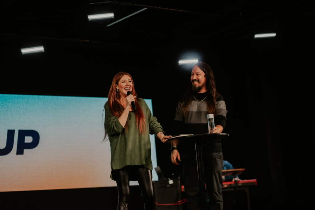 A woman with long red hair, wearing a green sweater and black leggings, speaks into a microphone next to a man with long dark hair, wearing a black and gray striped shirt. They stand by a round table with a glass of water, in front of a backdrop with a colorful screen.