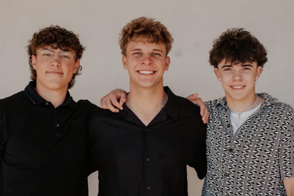 Three young men are standing closely together against a neutral background. The man on the left has curly hair and glasses, wearing a black shirt. The man in the middle has short hair and a black shirt, while the man on the right wears a patterned shirt.