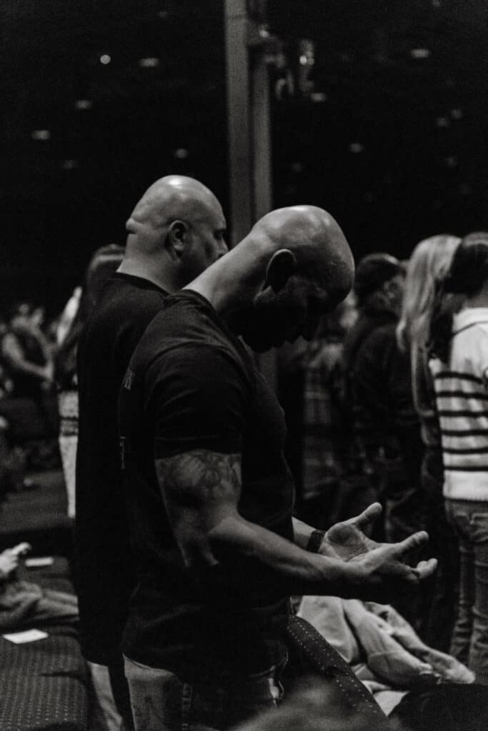 Two bald men stand among a crowd in a dimly lit setting. One man in a black shirt bows his head with his hands open, while the other man in a gray shirt stands beside him, both positioned in front of a group of people facing forward.