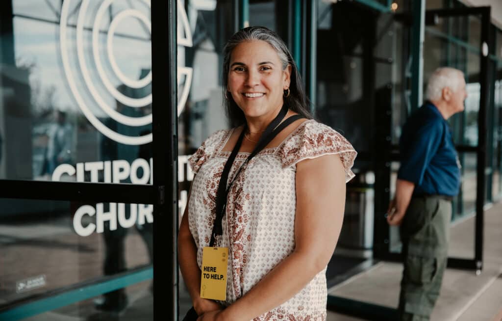 A woman with long hair wearing a patterned blouse stands in front of glass doors marked "CitiPoint Church." She has a badge reading "Here to Help" around her neck. In the background, a man is standing near a window.