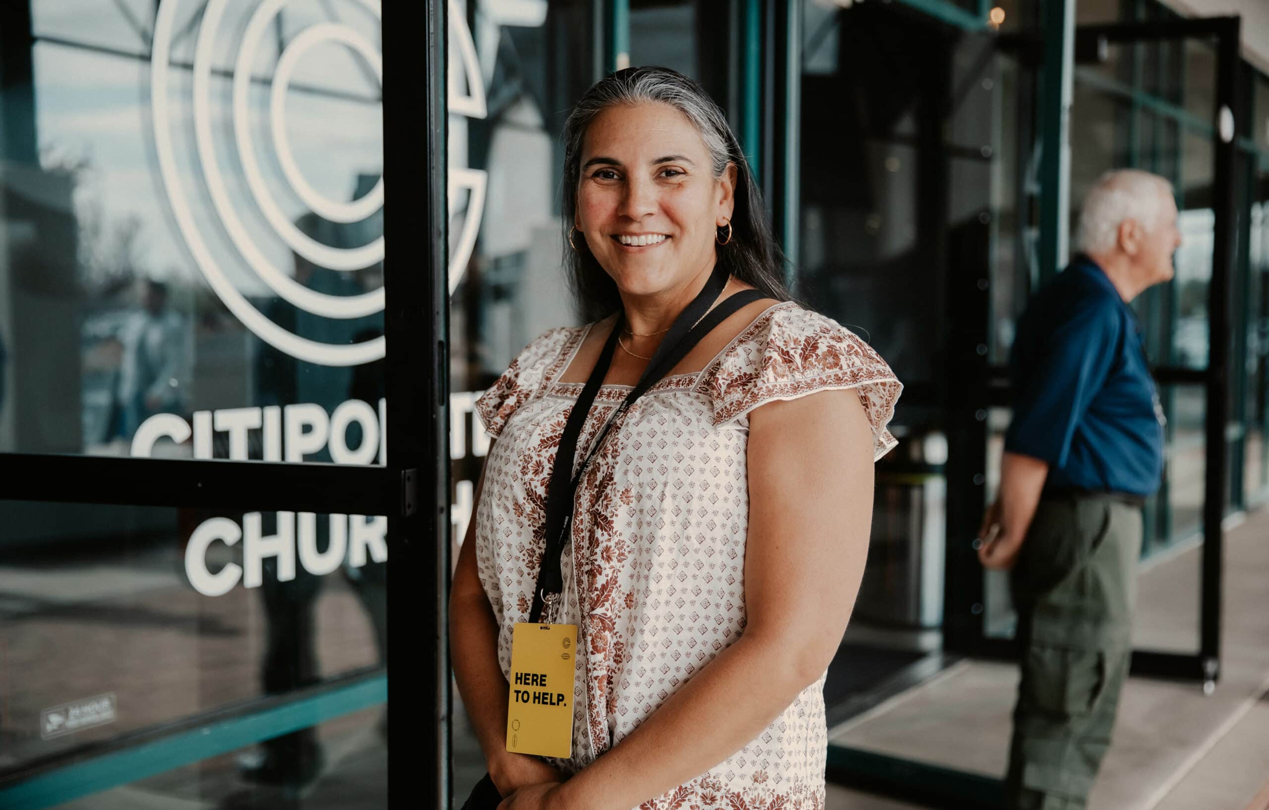 A woman with long hair wearing a patterned blouse stands in front of glass doors marked "CitiPoint Church." She has a badge reading "Here to Help" around her neck. In the background, a man is standing near a window.