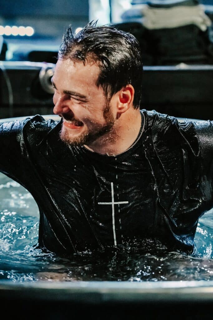 A man in a black t-shirt with a cross pattern is partially submerged in water, with his arms raised and a smile on his face. The setting appears to be a baptismal pool, with a blurred background of clothing and lighting.