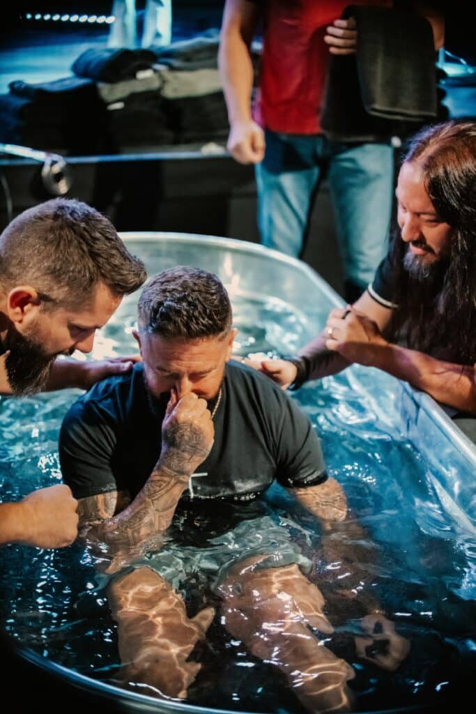 A man is seated in a metal tub filled with water, while two individuals, one on each side, support him. The scene appears to be in a dimly lit venue, with additional people visible in the background, engaged in the event.