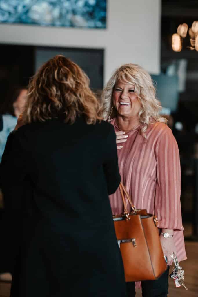 A woman with long, wavy blonde hair wearing a pink blouse stands in conversation, smiling at another woman whose back is turned. The setting is a well-lit indoor space with modern decor, including large windows in the background.