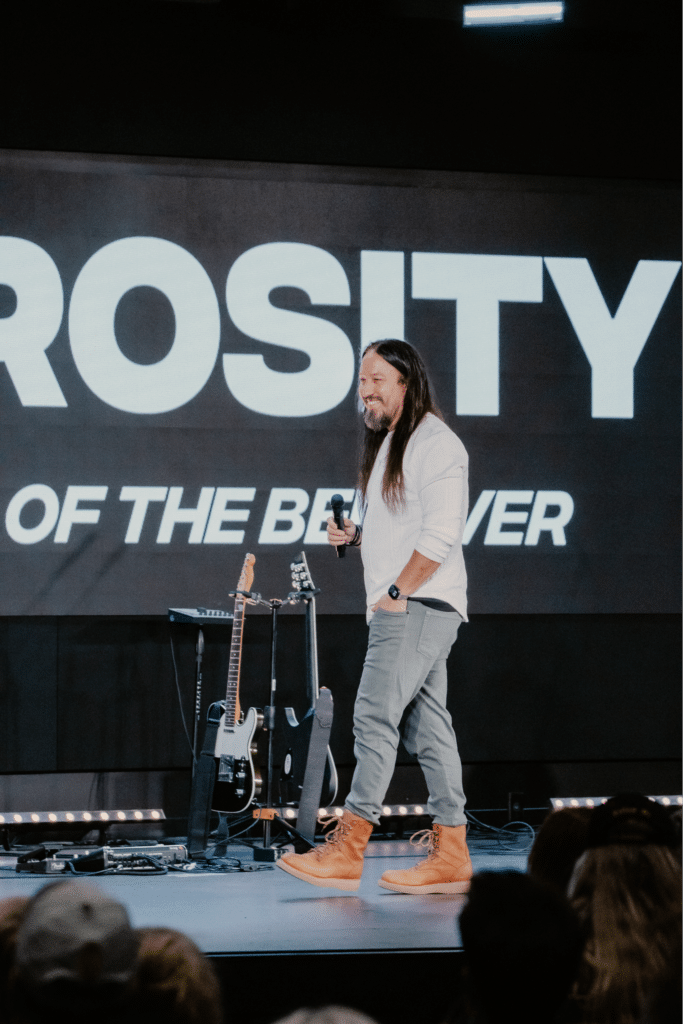 A man with long hair wearing a white long-sleeve shirt and gray pants is walking on stage. He is holding a microphone, and there is a guitar and an amplifier on the floor beside him. The background displays the word "GENEROSITY" in large letters.