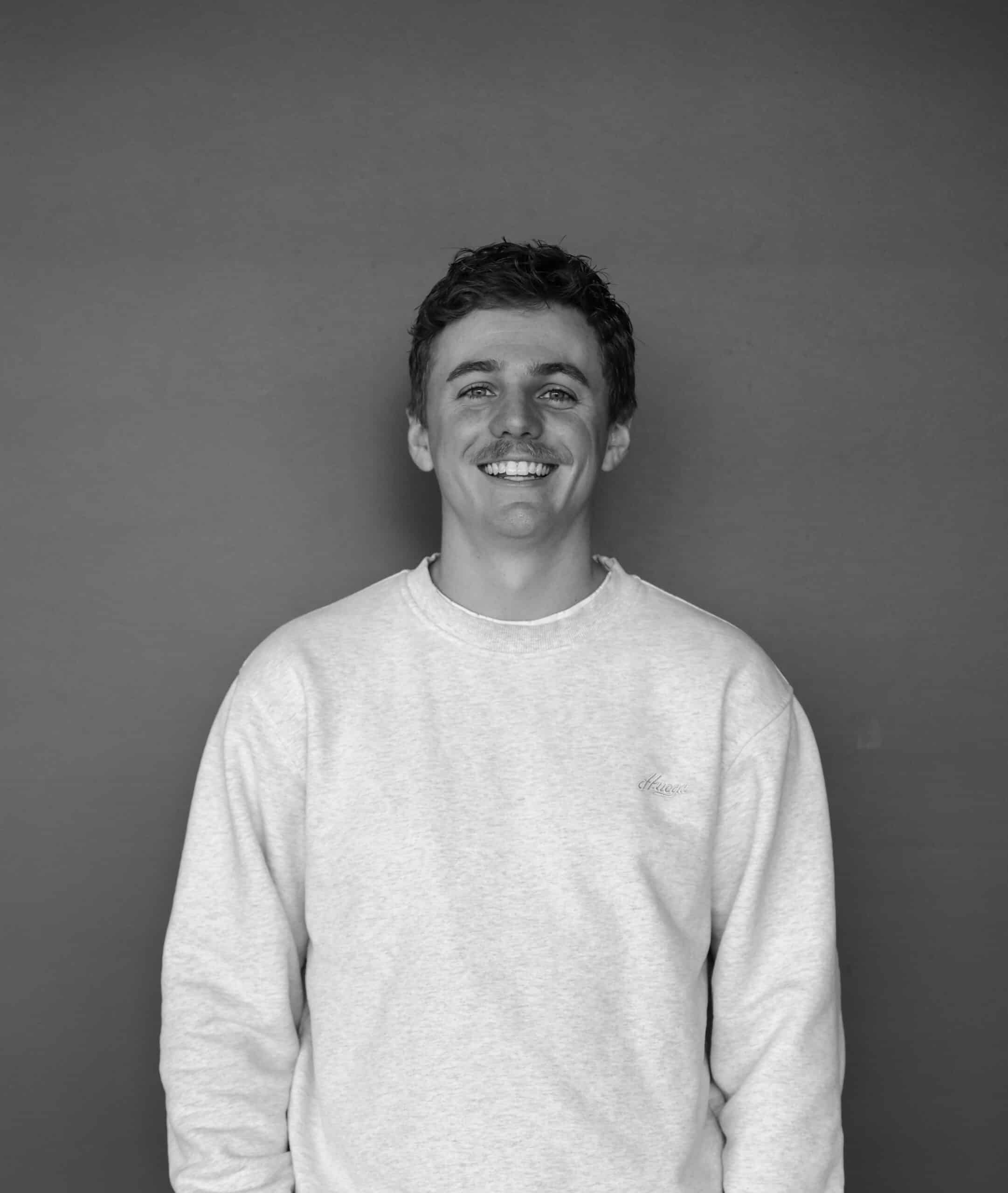 A young man with short dark hair and a mustache stands in front of a solid gray background. He is wearing a light gray sweatshirt and smiling directly at the camera.