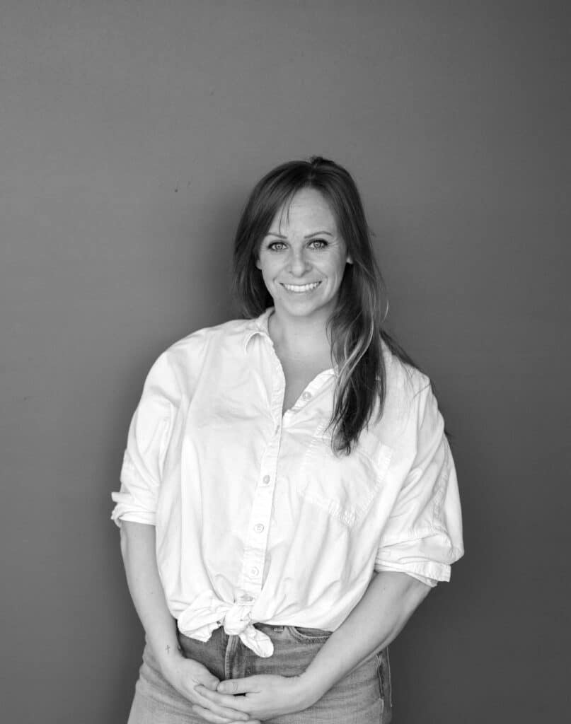 A woman stands against a gray background, wearing a white button-up shirt tied at the waist and high-waisted jeans. She has long hair and is posing with her hands clasped in front of her.