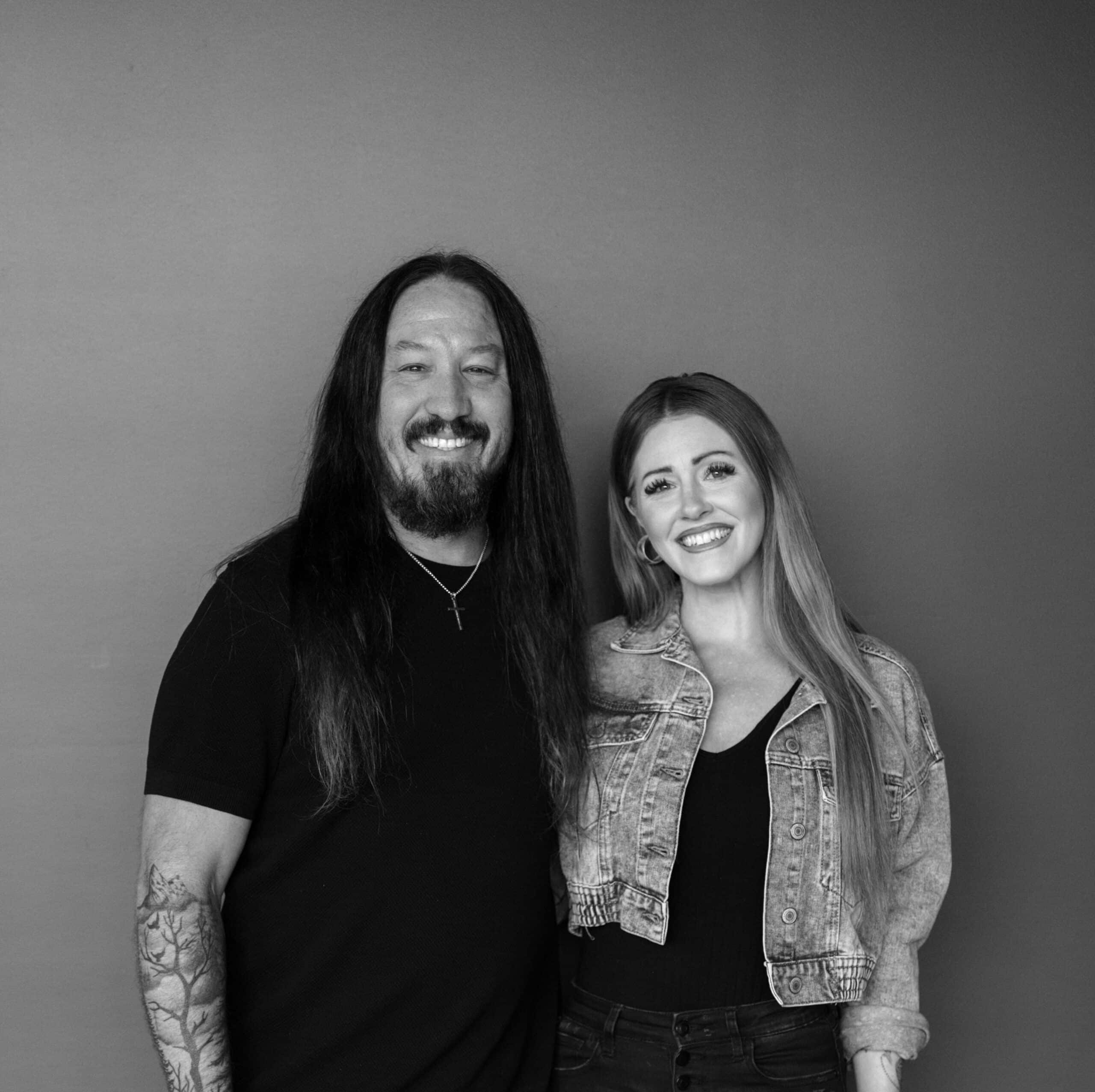 A man with long dark hair and a beard stands on the left, wearing a black shirt and silver necklace. A woman with long blonde hair, dressed in a black top and denim jacket, stands on the right. They are posing together in front of a gray background.