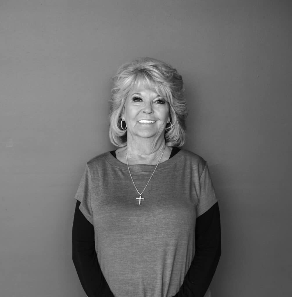 A woman with light curly hair and wearing a gray top over a black long-sleeve shirt stands with her hands clasped in front of her. She has large hoop earrings and a cross pendant necklace. The background is a solid light color.