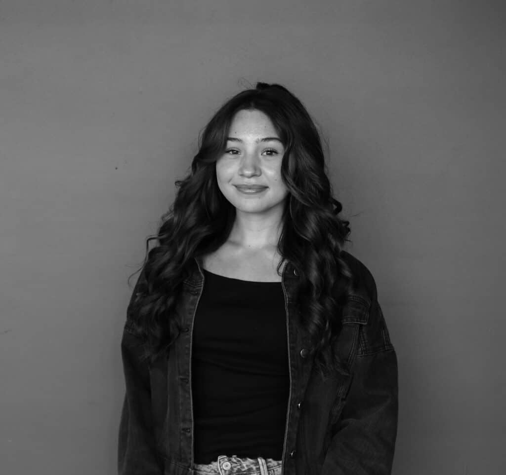 A young woman with long, wavy hair stands against a plain background, wearing a black top and an oversized denim jacket. The image is in black and white, showcasing her relaxed pose and the details of her outfit.
