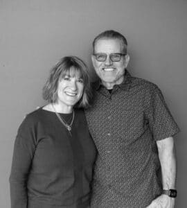 A woman with shoulder-length hair wearing a black sweater and layered necklaces stands next to a man with glasses and short hair, wearing a patterned button-up shirt. They are positioned against a neutral background.