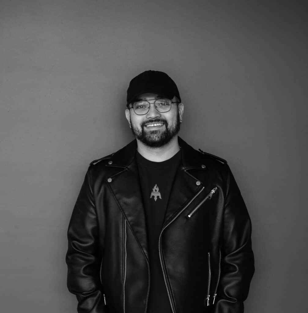 A man with glasses and a beard stands smiling against a gray background. He is wearing a black leather jacket over a dark t-shirt and a black cap.