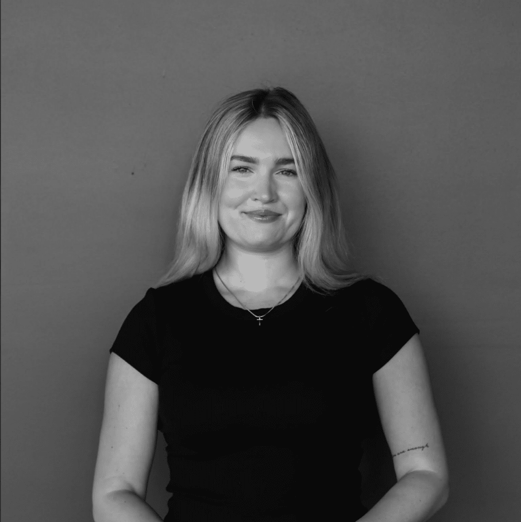 A woman with long, blonde hair is smiling while wearing a black t-shirt. She is seated against a neutral gray background, with her arms resting on her lap. A delicate necklace is visible around her neck.