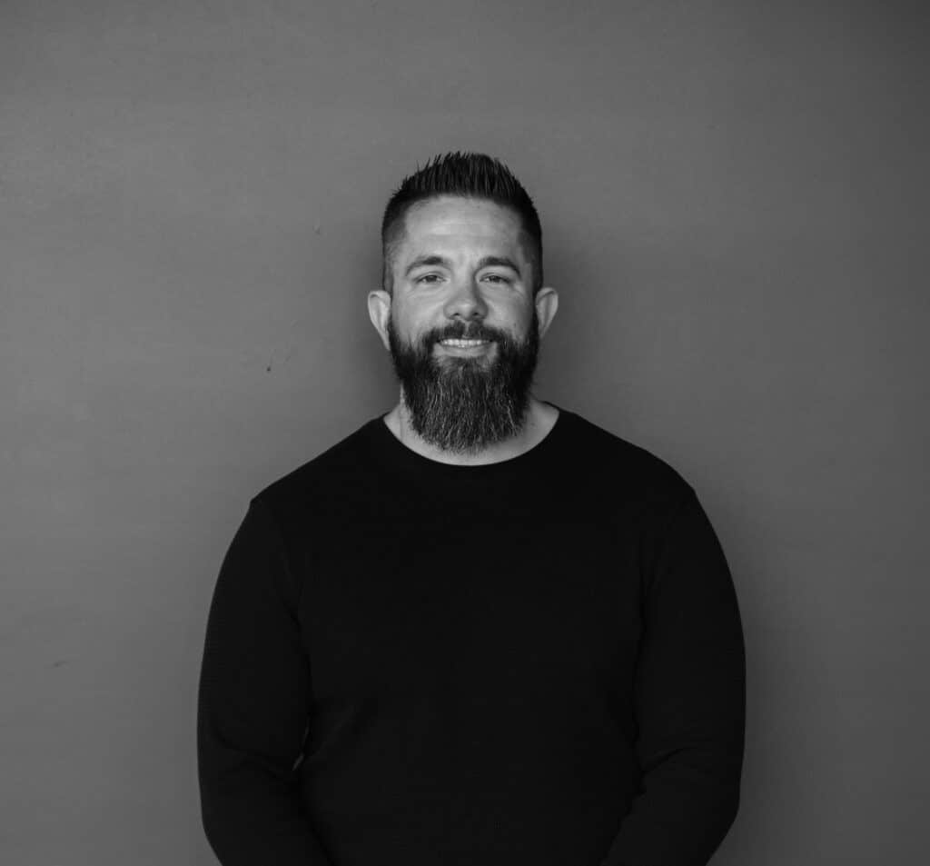 A man with a short beard and styled hair stands in front of a plain gray background. He is wearing a black long-sleeve shirt and facing the camera directly.