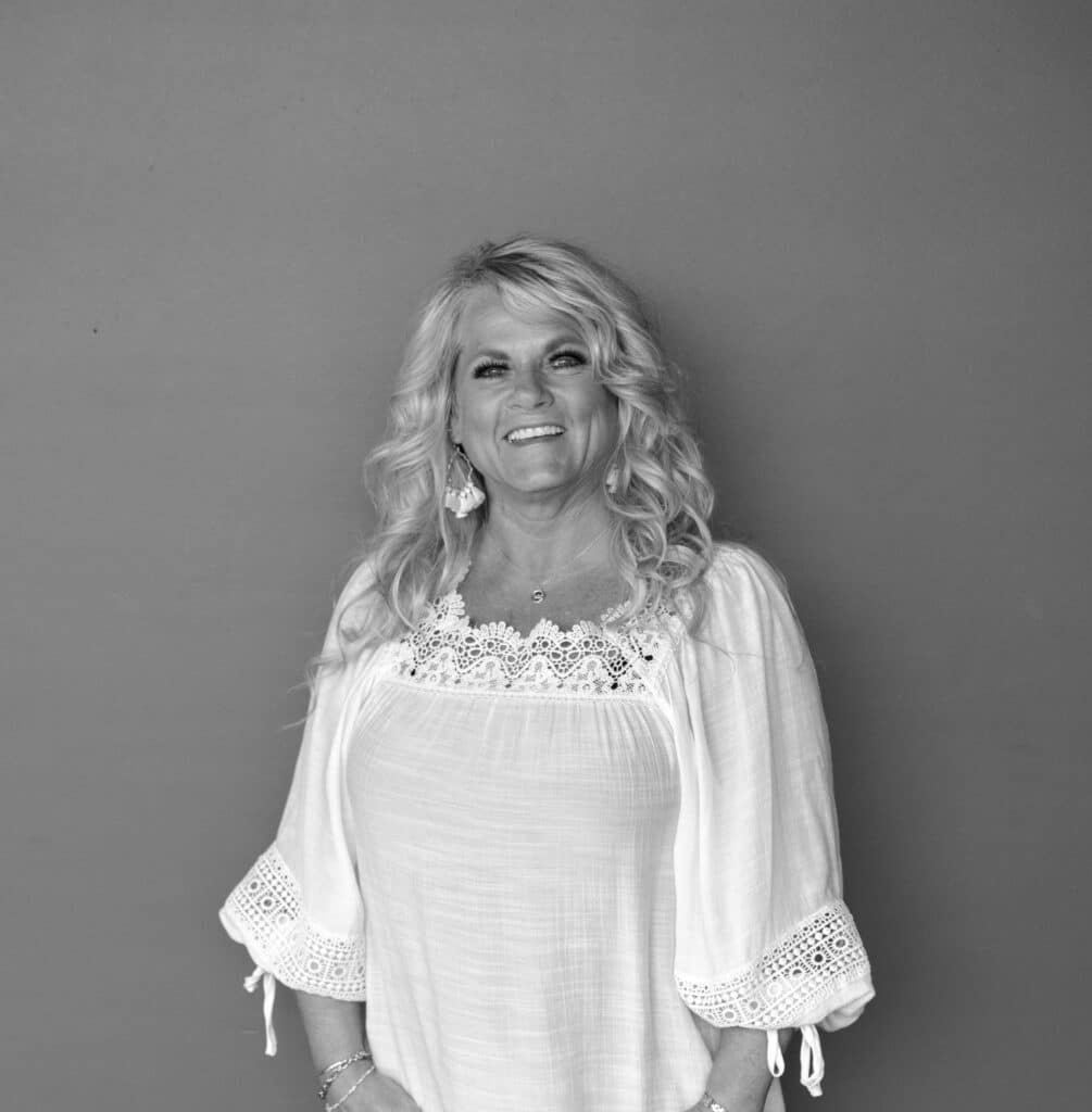A woman with long, wavy blonde hair is standing against a plain gray background. She is wearing a white blouse with lace detailing on the collar and sleeves. The image is in black and white.